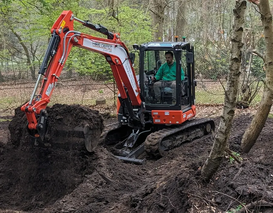 A man operates an excavator, digging a deep hole in the ground under clear blue skies.