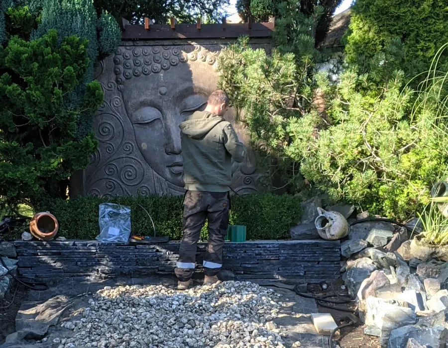 A man meticulously carves a stone sculpture in a serene garden setting, surrounded by greenery and natural beauty.