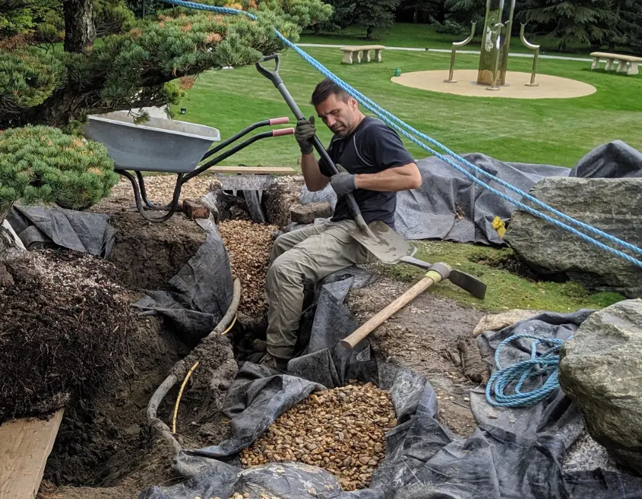 A man tends to a garden, arranging gravel and nurturing various plants in a serene outdoor setting.